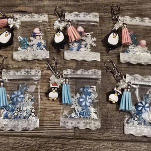 May include: Keychains featuring a penguin charm, tassel, and a clear bag filled with blue and white decorative elements. The keychains have silver-colored hardware and a snowflake-patterned border. Some penguins wear Santa hats.