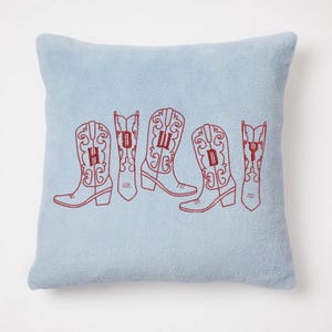 May include: A light blue square pillow with embroidered red cowboy boots spelling out "HOWDY". The boots are detailed with decorative stitching. The pillow has a soft, textured appearance, suggesting a cozy home decor item.