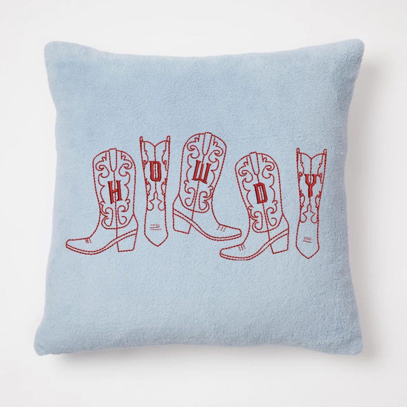 May include: A light blue square pillow with embroidered red cowboy boots spelling out "HOWDY". The boots are detailed with decorative stitching. The pillow has a soft, textured appearance, suggesting a cozy home decor item.