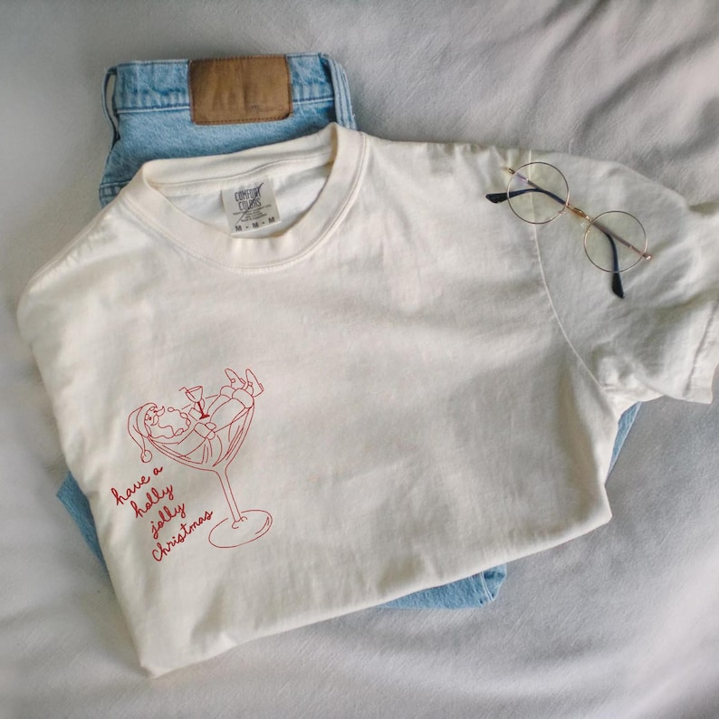 Op de afbeelding: Cr&egrave;mekleurig T-shirt met een rode afbeelding van de kerstman in een martini glas, met de tekst "have a holly jolly Christmas". Het shirt wordt gecombineerd met een lichtblauwe jeans en een ronde bril.