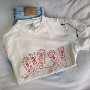 May include: Cream-colored t-shirt with the word "HOWDY" embroidered in red on the front, featuring stylized cowboy boot designs. The shirt is paired with light blue denim jeans and round-framed glasses.