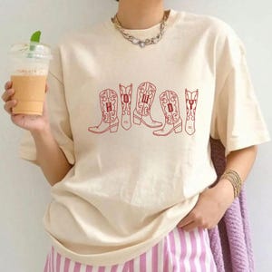 May include: Cream-colored t-shirt featuring a red graphic of five cowboy boots. Each boot has a letter inside, spelling out a word. The shirt has short sleeves and a relaxed fit. The person is wearing a gold necklace and bracelets, and pink and white striped pants.