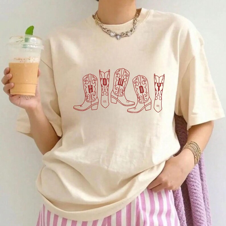 May include: Cream-colored t-shirt featuring a red graphic of five cowboy boots. Each boot has a letter inside, spelling out a word. The shirt has short sleeves and a relaxed fit. The person is wearing a gold necklace and bracelets, and pink and white striped pants.