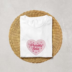 May include: White t-shirt with a pink heart-shaped embroidered design. The heart has a scalloped edge and the words "Probably Smut" in pink script. Small red hearts are scattered around the heart design.