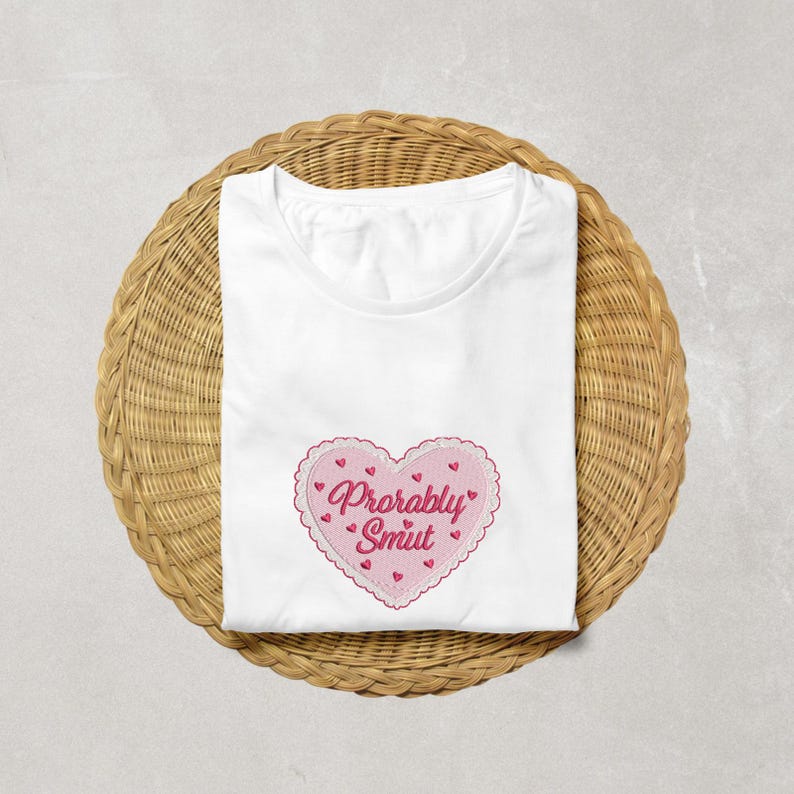 May include: White t-shirt with a pink heart-shaped embroidered design. The heart has a scalloped edge and the words "Probably Smut" in pink script. Small red hearts are scattered around the heart design.