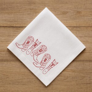 May include: White square napkin with a red embroidered design of five cowboy boots. The boots are arranged diagonally across the napkin, with intricate detailing. The napkin is made of a soft, likely linen or cotton material.