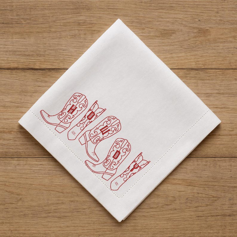 May include: White square napkin with a red embroidered design of five cowboy boots. The boots are arranged diagonally across the napkin, with intricate detailing. The napkin is made of a soft, likely linen or cotton material.