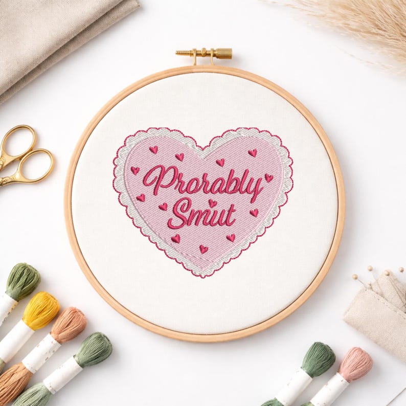 May include: A pink heart-shaped embroidery hoop with the words "Probably Smut" in a cursive font. The heart is decorated with small red hearts and a scalloped edge. The wooden hoop is surrounded by embroidery supplies.
