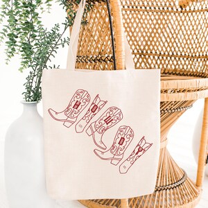 May include: Cream-colored tote bag with a red embroidered design of cowboy boots spelling out the word "HOLD" in a stylized font. The bag is hanging on a wicker chair, with a white vase and greenery in the background.