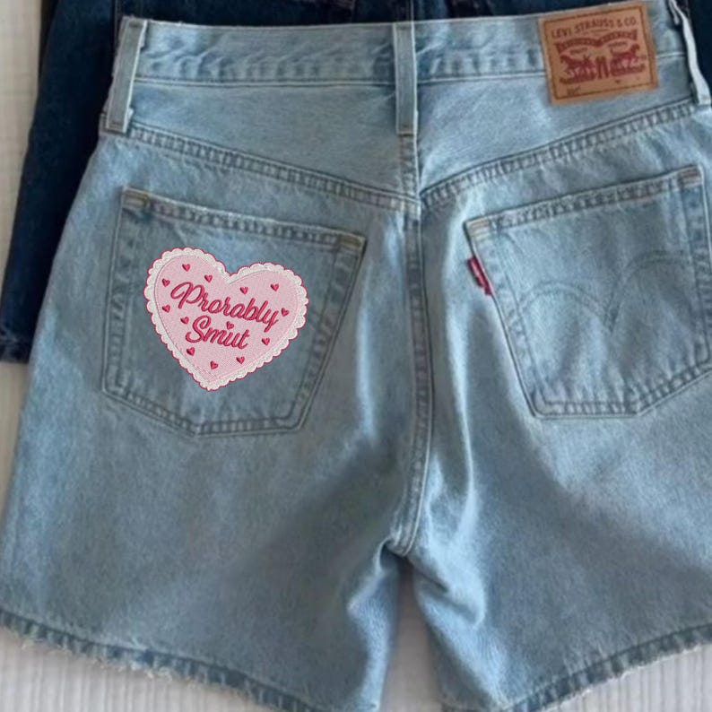 May include: Light blue denim shorts with a heart-shaped patch on the back pocket. The patch is pink with the words "Probably Smut" embroidered in a cursive font. The shorts have a classic five-pocket design and a red Levi's tag.