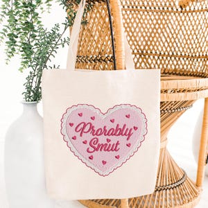 May include: Cream-colored tote bag with a pink heart design. The heart is embroidered with the words "Probably Smut" in a cursive font, surrounded by small pink hearts. The bag has two cream-colored handles.