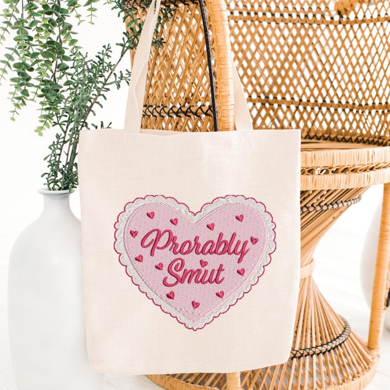 May include: Cream-colored tote bag with a pink heart design. The heart is embroidered with the words "Probably Smut" in a cursive font, surrounded by small pink hearts. The bag has two cream-colored handles.