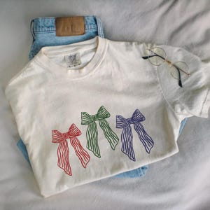 以下が含まれることがあります： 赤、緑、青の3色のリボンデザインが施されたクリーム色のTシャツ。明るいブルーのジーンズと丸いフレームのメガネと組み合わせています。Tシャツはクルーネックで半袖です。