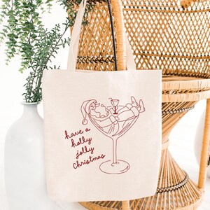 Op de afbeelding: Off-white canvas draagtas met een rode lijntekening van de Kerstman die ontspant in een cocktailglas. De tekst "have a holly jolly Christmas" is in rode letters geschreven. De tas heeft twee beige handvatten.