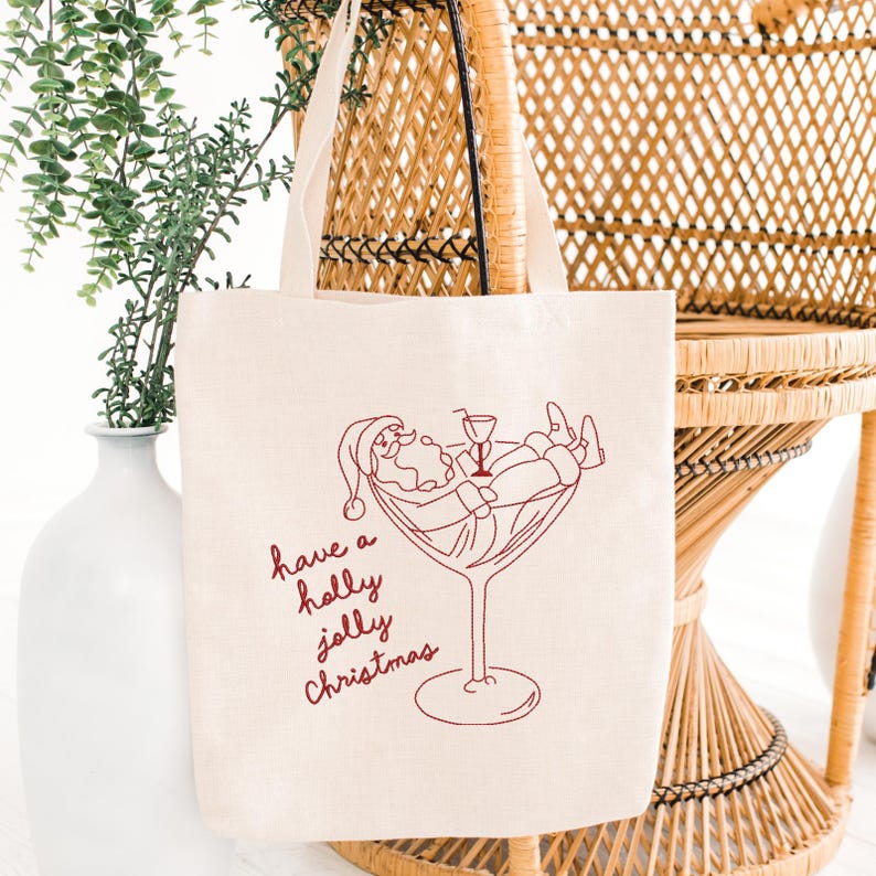 Op de afbeelding: Off-white canvas draagtas met een rode lijntekening van de Kerstman die ontspant in een cocktailglas. De tekst "have a holly jolly Christmas" is in rode letters geschreven. De tas heeft twee beige handvatten.