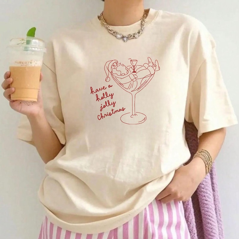 Op de afbeelding: Cr&egrave;mekleurig T-shirt met een rode afbeelding van de Kerstman die ontspant in een cocktailglas, met de tekst "have a holly jolly Christmas". Het shirt heeft korte mouwen en een relaxte pasvorm.