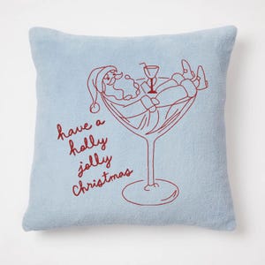 Op de afbeelding: Een lichtblauw vierkant kussen met een rood geborduurd ontwerp. Het ontwerp toont de kerstman die ontspant in een martiniglas, met de tekst "have a holly jolly Christmas" in rode letters.