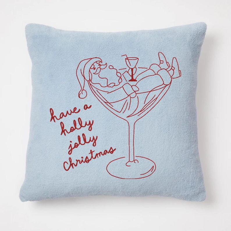 Op de afbeelding: Een lichtblauw vierkant kussen met een rood geborduurd ontwerp. Het ontwerp toont de kerstman die ontspant in een martiniglas, met de tekst "have a holly jolly Christmas" in rode letters.