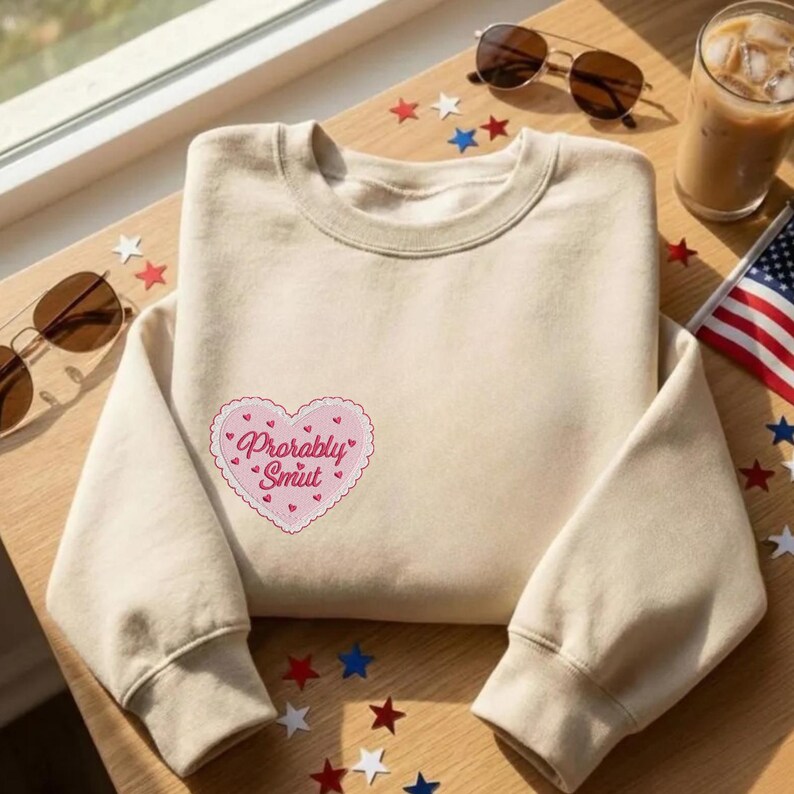 May include: A beige sweatshirt with a pink heart-shaped patch that reads "Probably Smut" in cursive. The sweatshirt is folded on a wooden surface, with sunglasses, a drink, and an American flag in the background.