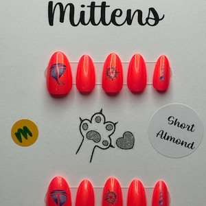 May include: Vibrant orange press-on nails with nautical and hot air balloon designs. The nails are almond-shaped and branded as "Murder Mittens." The text "Atomic Polish - Shantay, You Stay" is also visible on the image.