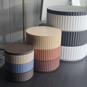 May include: A collection of decorative round boxes in various colors, including brown, beige, blue, and white. Each box has a ribbed design on the sides and a flat, circular lid. The boxes are stacked and arranged on a dark surface.