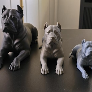 Könnte beinhalten: Drei Hundefiguren in absteigender Größe, jede in liegender Position mit aufmerksamen Ohren und nach vorne gerichtetem Blick. Die größte ist mattschwarz, die mittlere hellgrau und die kleinste etwas dunkler grau, alle mit glatter Oberfläche.