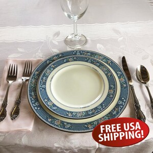 May include: A set of white dinnerware with a blue and floral pattern. The set includes a dinner plate, a salad plate, a fork, a knife, and a spoon. The plates have a gold trim. The tableware is set on a white tablecloth with a lace pattern.