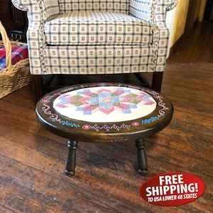 May include: A round wooden stool with a quilted fabric top. The stool has three legs and is painted with pink, blue, and green accents. The fabric top features a patchwork design with pink, green, and blue squares.