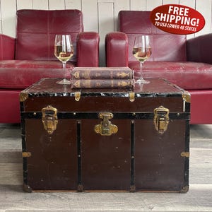 May include: A dark brown vintage trunk with black trim and brass-toned metal latches, featuring two stacked antique books and two clear glass wine glasses filled with white liquid placed on its flat top surface in front of two red leather chairs.