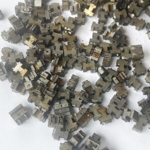 May include: A pile of small, weathered, metallic letterpress printing type pieces. The individual pieces are mostly gray with some showing signs of rust. The shapes are primarily T-shaped, with some showing other letter forms.