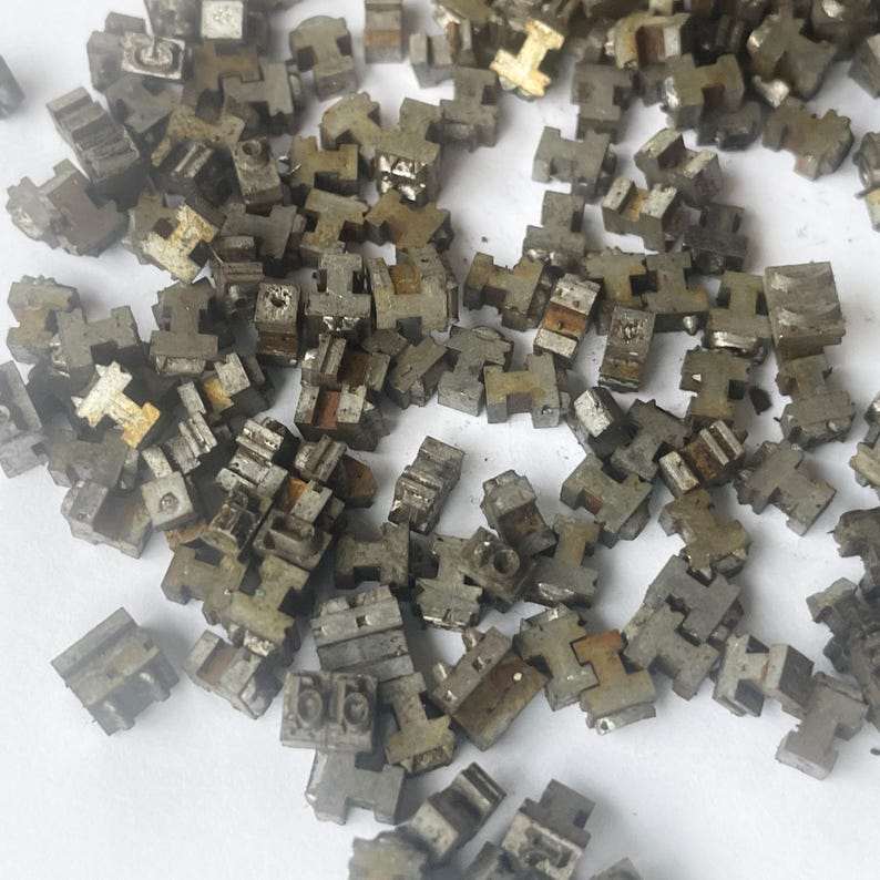 May include: A pile of small, weathered, metallic letterpress printing type pieces. The individual pieces are mostly gray with some showing signs of rust. The shapes are primarily T-shaped, with some showing other letter forms.