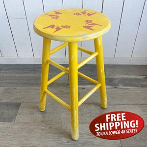 May include: A yellow wooden stool with a round seat featuring four red horse-shaped motifs. The stool has four legs connected by horizontal support rungs, and the yellow paint shows signs of wear and distress throughout the entire structure.