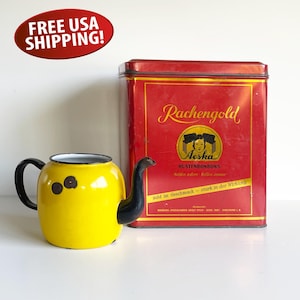 May include: A yellow enamel teapot with a black handle and spout, next to a red rectangular tin with gold lettering and the word "Aeska". The teapot has three brown spots on the side.