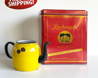 May include: A yellow enamel teapot with a black handle and spout, next to a red rectangular tin with gold lettering and the word "Aeska". The teapot has three brown spots on the side.