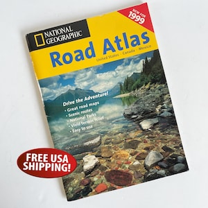 Puede incluir: Un Atlas de Carreteras National Geographic de 1999. La portada presenta un paisaje escénico con montañas y un lago. El título está en letras grandes azules y amarillas. Incluye el texto "Drive the Adventure!" y "United States Canada Mexico".