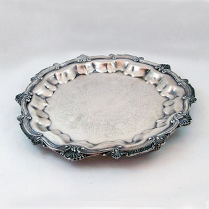 May include: A silver serving tray with a scalloped edge and an ornate design. The tray has a raised rim and a smooth, reflective surface.