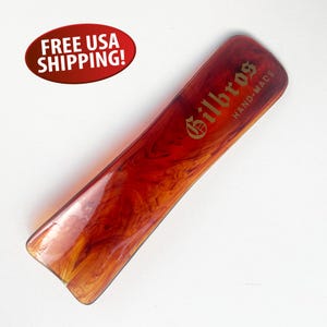 May include: A rectangular, hand-made comb with a marbled red and orange pattern. The comb has the word "Gilbros" in gold lettering, with "HAND-MADE" below it. The comb is on a white background.