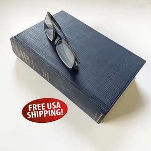 May include: A navy blue hardcover book with the title "The Book of English Romantic Poetry" and black-framed eyeglasses resting on top. The book has a textured cover and a signature in the upper right corner.