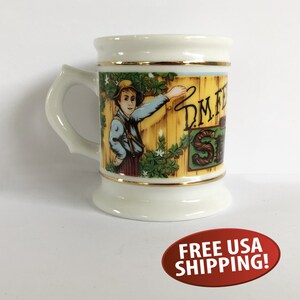 May include: White ceramic mug with a gold rim and a colorful illustration of a boy pointing to a sign that reads "D.M.F.  Strawberry Shortcake".