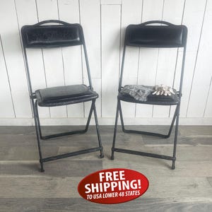 May include: Two black folding chairs with padded seats and backs. The chairs have a metal frame and are set against a white wooden wall. One chair has a gray scarf and gloves on the seat.