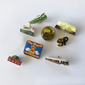May include: A collection of nine enamel pins featuring various transportation themes. The pins are colorful and feature images of trains, a train station, and a train track. The pins are all different sizes and shapes.