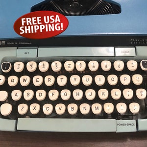 May include: A light blue vintage typewriter with a black keyboard and white keys. The typewriter has a clear plastic cover over the keys. The words "FREE USA SHIPPING!" are printed on a red oval sticker on the top of the typewriter. The words "Smith-Corona" are printed on the front of the typewriter. The words "Galaxy 12" are printed on the right side of the typewriter.