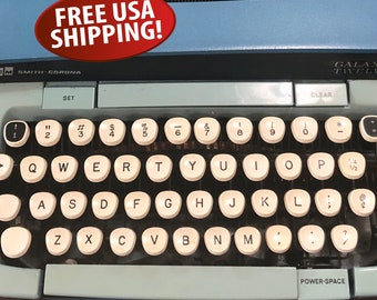 May include: A light blue Smith-Corona Galaxy Twelve typewriter featuring a QWERTY keyboard layout with off-white circular keys. The machine includes a power-space bar, a set key, and a clear key, with black text printed on the frame.