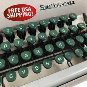 May include: A vintage Smith Corona typewriter with a green keyboard and white keys. The keys are labeled with letters, numbers, and symbols. The typewriter is in good condition and has a classic look.