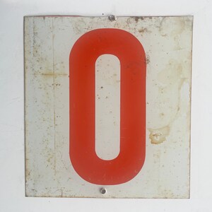 May include: Vintage metal sign featuring a bold red number zero on a white background. The sign has a rectangular shape with visible signs of wear and tear, including rust and discoloration, adding to its antique charm. Two rivets secure the sign.