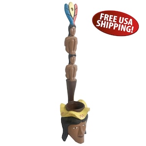 May include: A hand-carved wooden sculpture featuring a Native American figure with a headdress and two seated figures stacked above. The sculpture has a brown, yellow, and blue color scheme. The sculpture is approximately 12 inches tall.