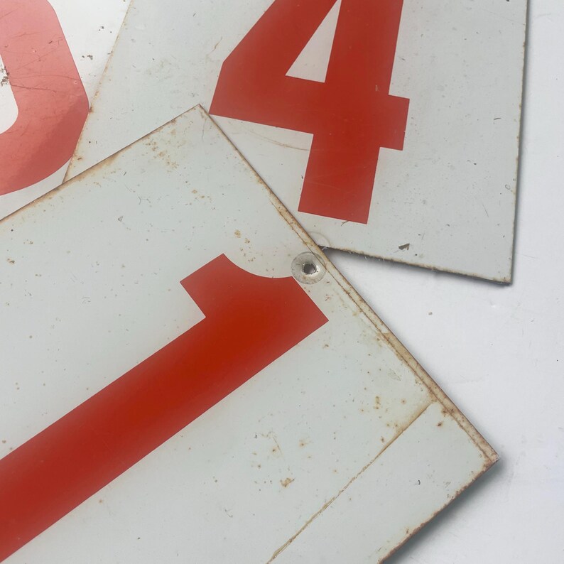 May include: Close-up of vintage metal number signs. The signs are white with large red numbers, including the number 1 and the number 4. The signs show signs of wear and tear, with rust and scratches visible on the surface.