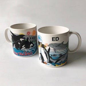 May include: Two white ceramic mugs with colorful illustrations of sea life. One mug features a penguin, a dolphin, and a shark. The other mug features a group of orcas. Both mugs have the text "Sea World" and "ED" printed on them.