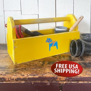 May include: A bright yellow wooden tool caddy featuring a blue Dala horse graphic on the front panel. The rectangular carrier has a sturdy top handle and contains various gardening tools and black plastic pots inside the open storage compartment.