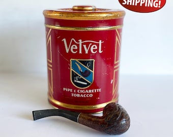 Può includere: Una scatola rossa di tabacco Velvet con finiture dorate e un logo raffigurante una pipa e una sigaretta. Una pipa di tabacco marrone intagliata è posta davanti alla scatola. La scatola recita "Velvet Pipe & Cigarette Tobacco."